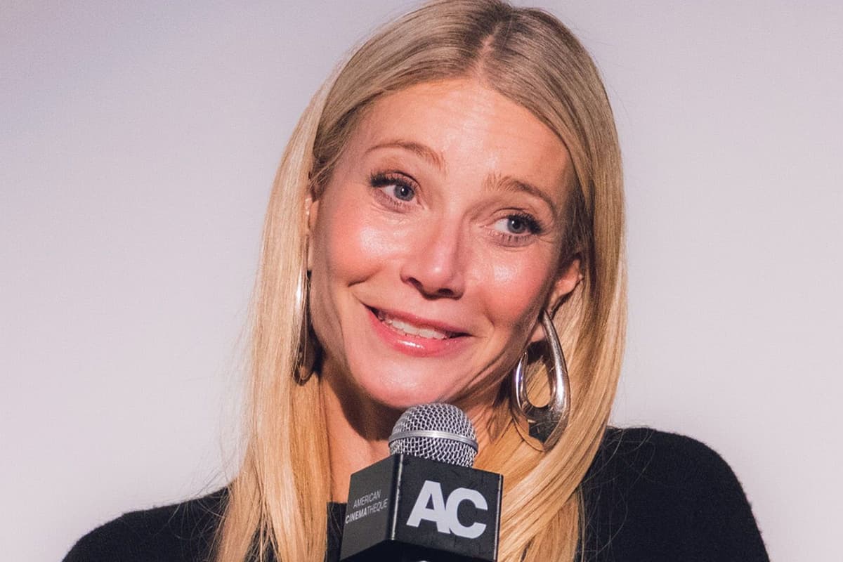 Smiling blonde woman in a black sweater and hoop earrings, holding a microphone against a plain light grey backdrop