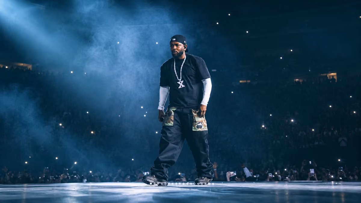 Kendrick Lamar walking across a stadium stage holding a microphone, with phone lights in the crowd behind him