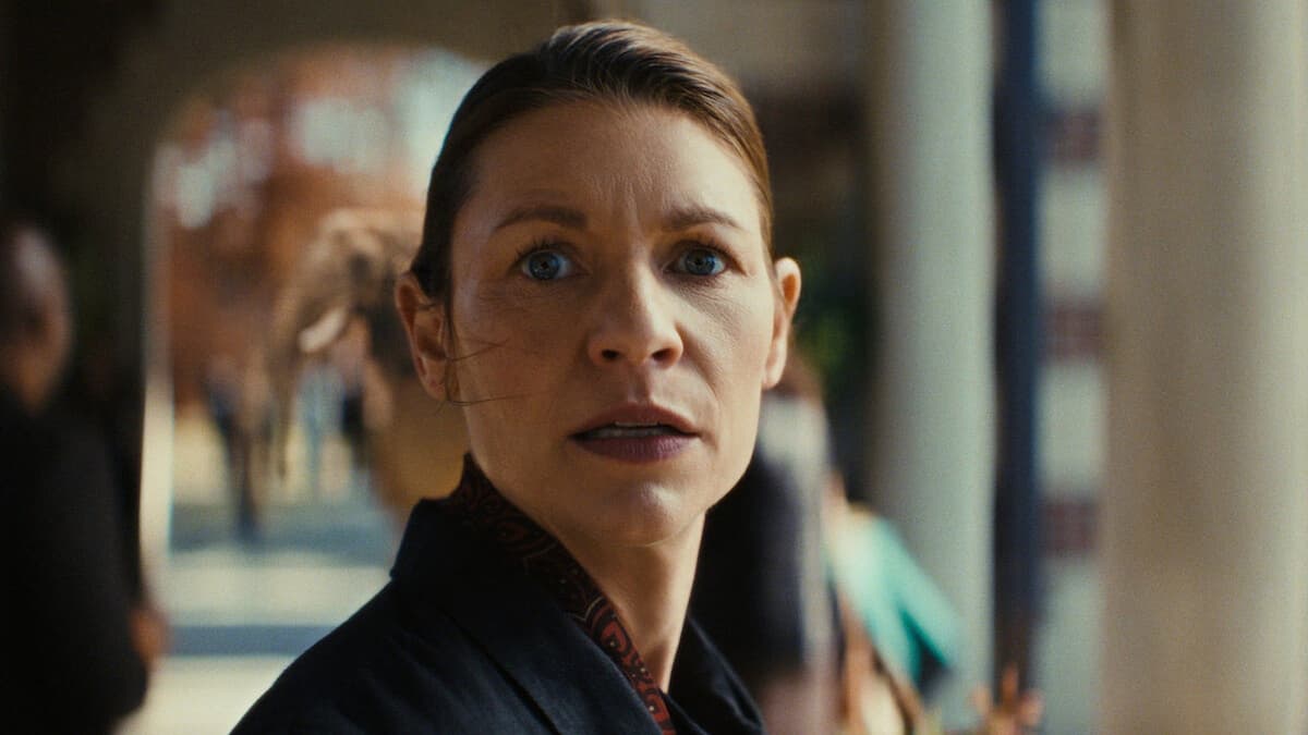 Close-up of a woman with slicked-back hair and blue eyes, looking startled in a dark jacket under an arcade