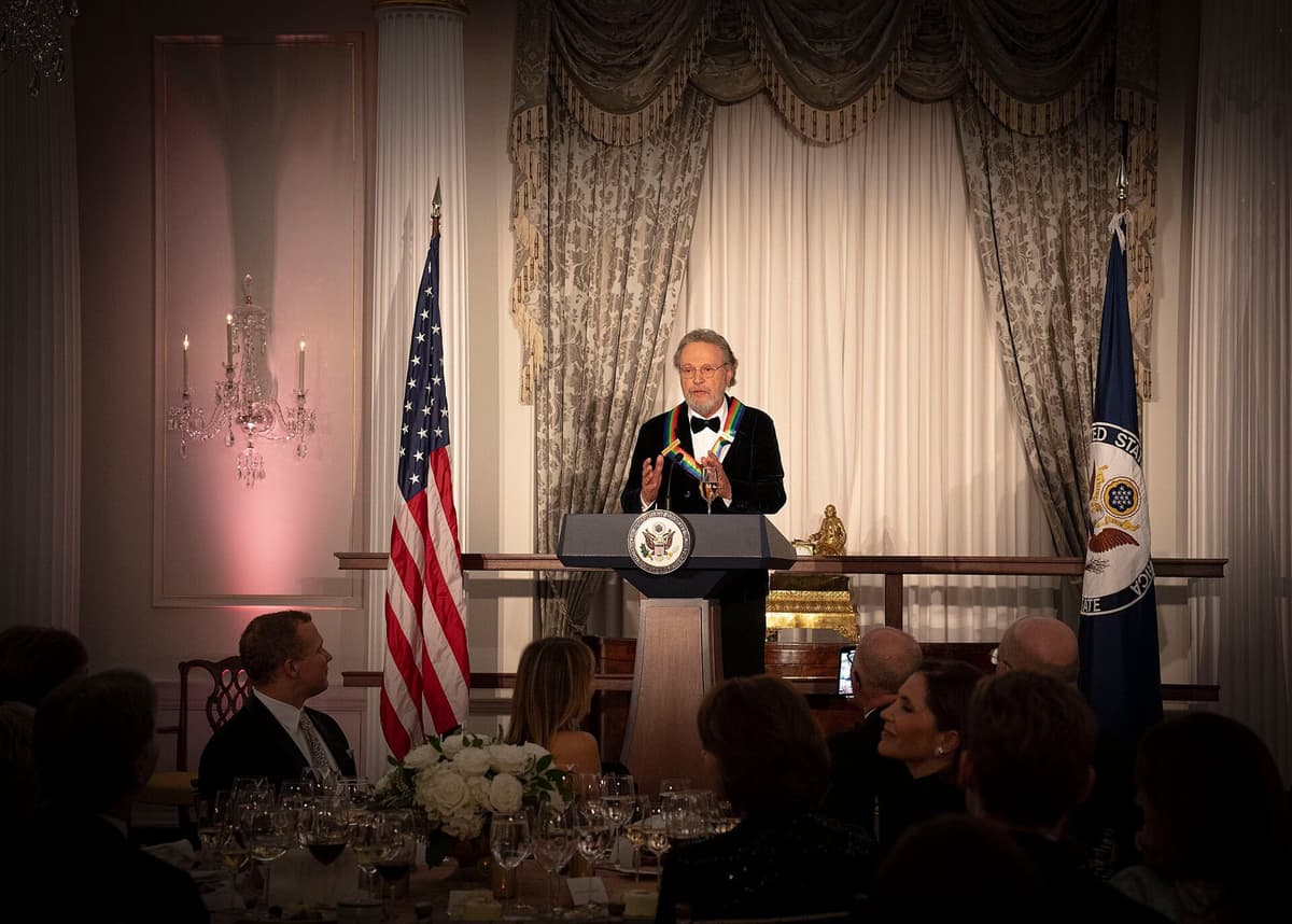 Billy Crystal in a tuxedo with a rainbow honors medallion speaks at a lectern to a seated dinner audience in a formal room.