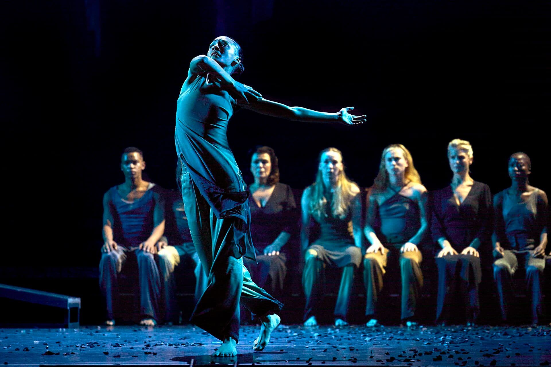 A dancer performs on stage with ensemble members seated in the background.