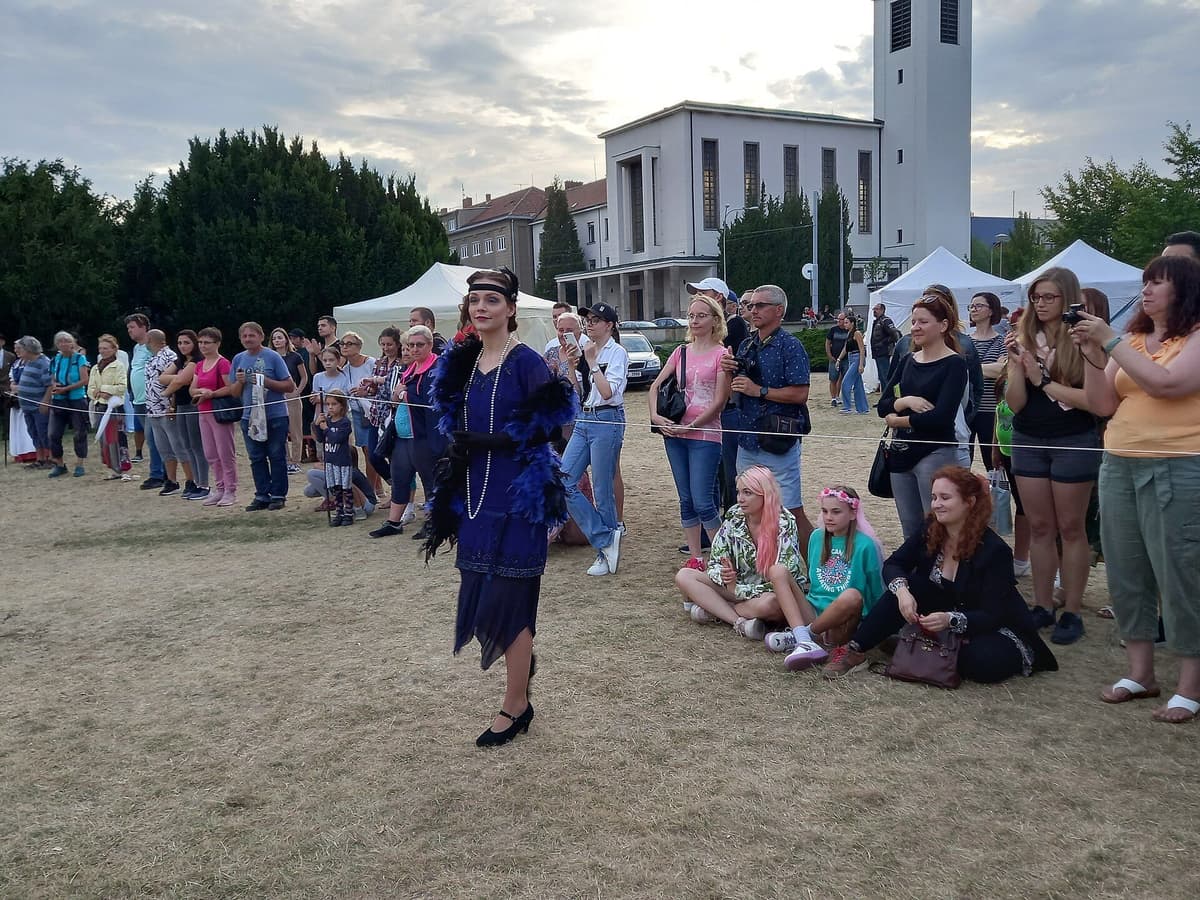 A woman in a blue flapper-style dress and feather boa walks on grass as spectators watch behind a rope line