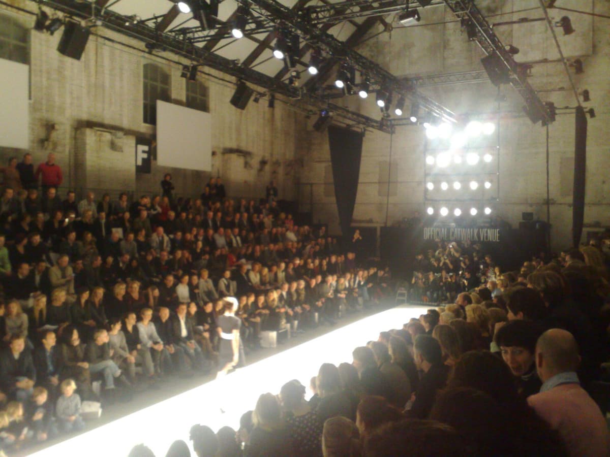 Brightly lit catwalk in a industrial hall with a model walking and a packed audience on both sides
