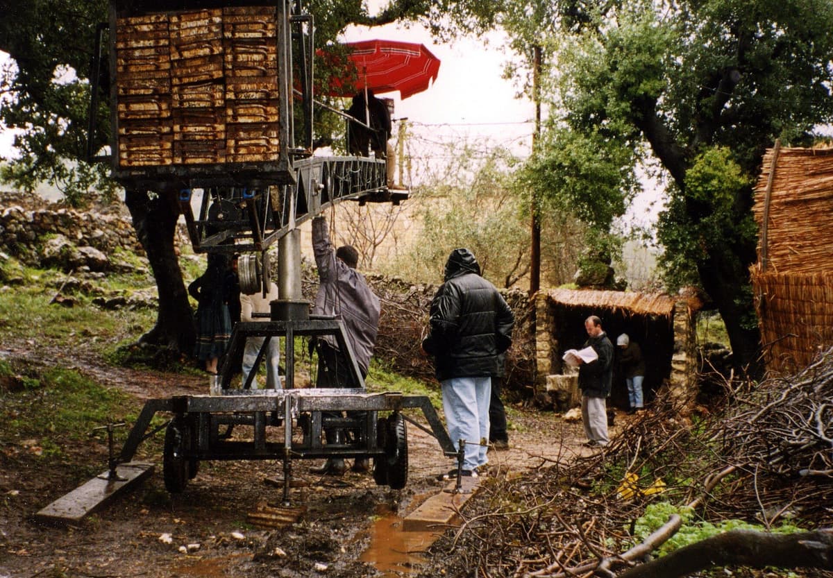 Film crew on a wet wooded location beside a camera crane on a wheeled platform, with a person reading pages