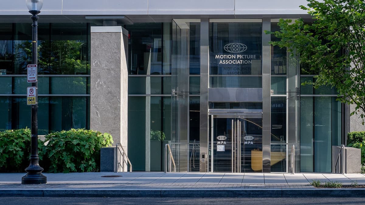 Glass entrance of a office building with the Motion Picture Association name and logo on the facade, beside a street lamp and shrubs.