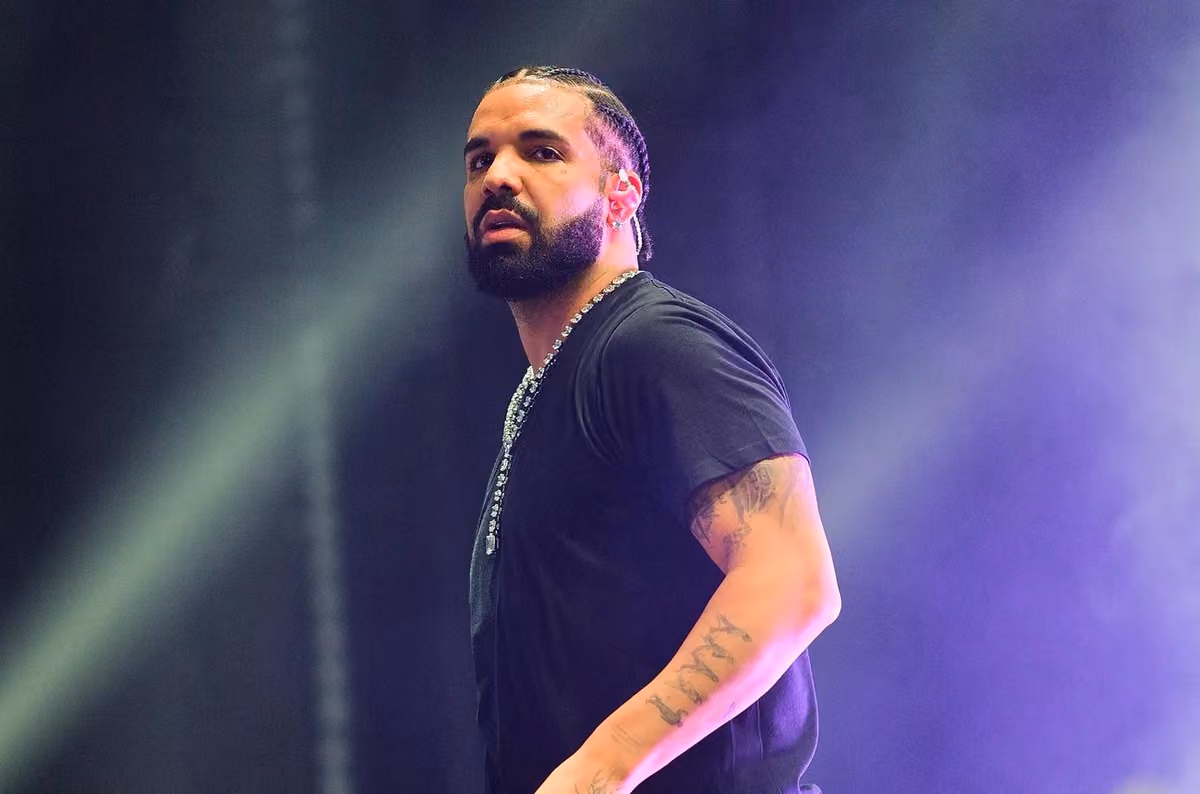 Drake performing on stage in a black T-shirt and diamond chain under purple stage lights