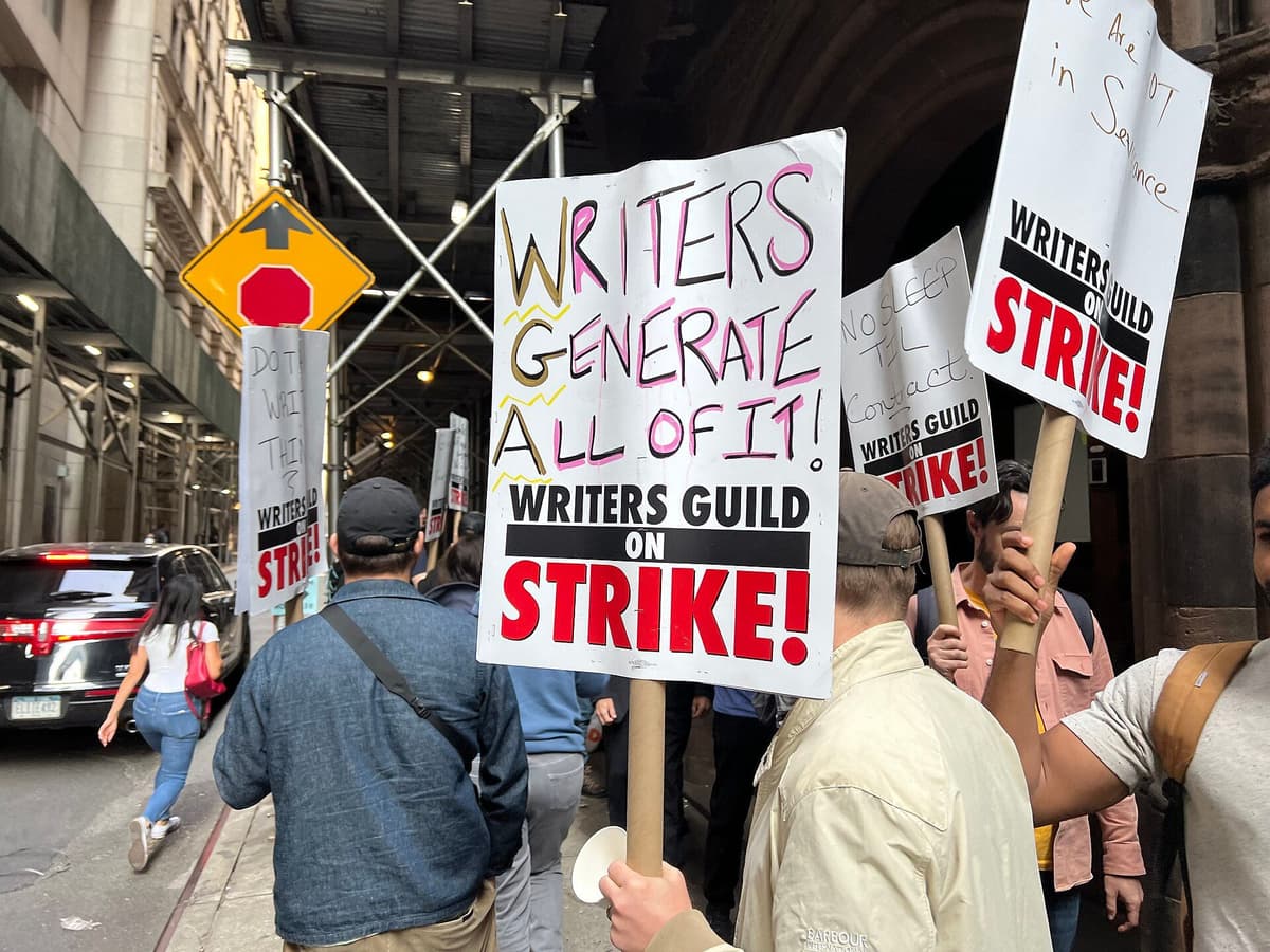 Striking writers walk a picket line on a city sidewalk; one sign reads 'Writers Generate All of It!'