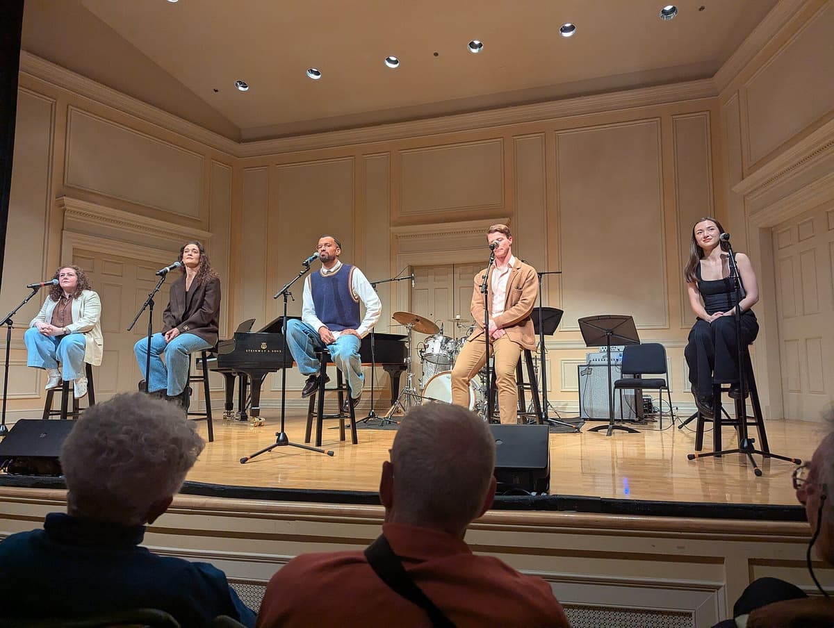 Five performers seated on stools with microphones on a wood-floored stage, with a piano and drum kit, as audience watch