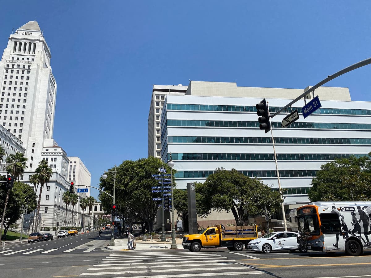 Downtown Los Angeles street intersection with traffic, a Metro bus, and City Hall tower on the left under blue sky.