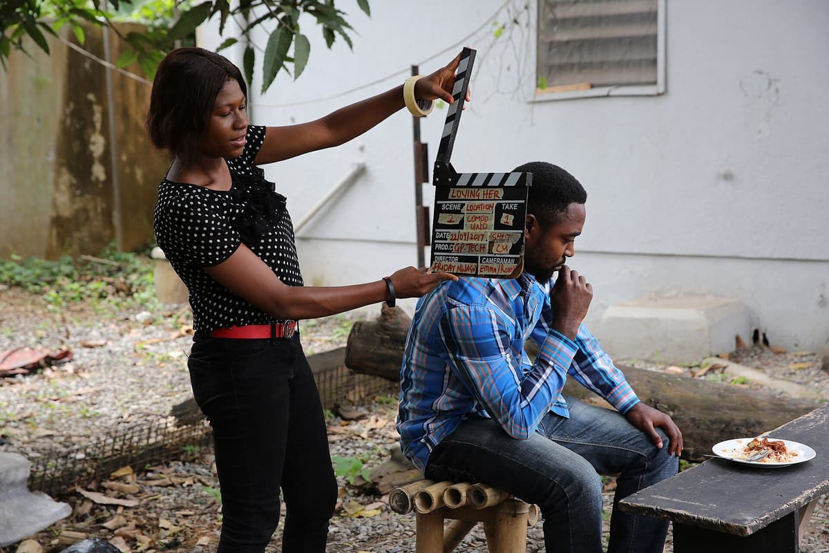 A crew member holds a clapperboard in front of a seated actor on an outdoor film set.