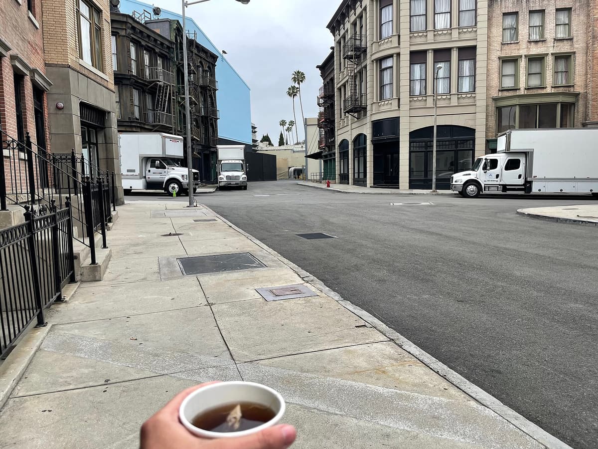 Paved street on a studio backlot with building facades and white trucks, a hand holding a cup of tea in front