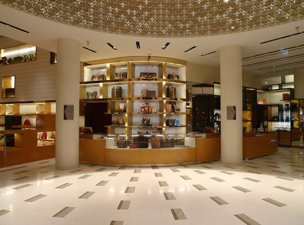 Boutique interior with handbags and trunks on lit gold-framed shelves, round columns and a patterned ceiling