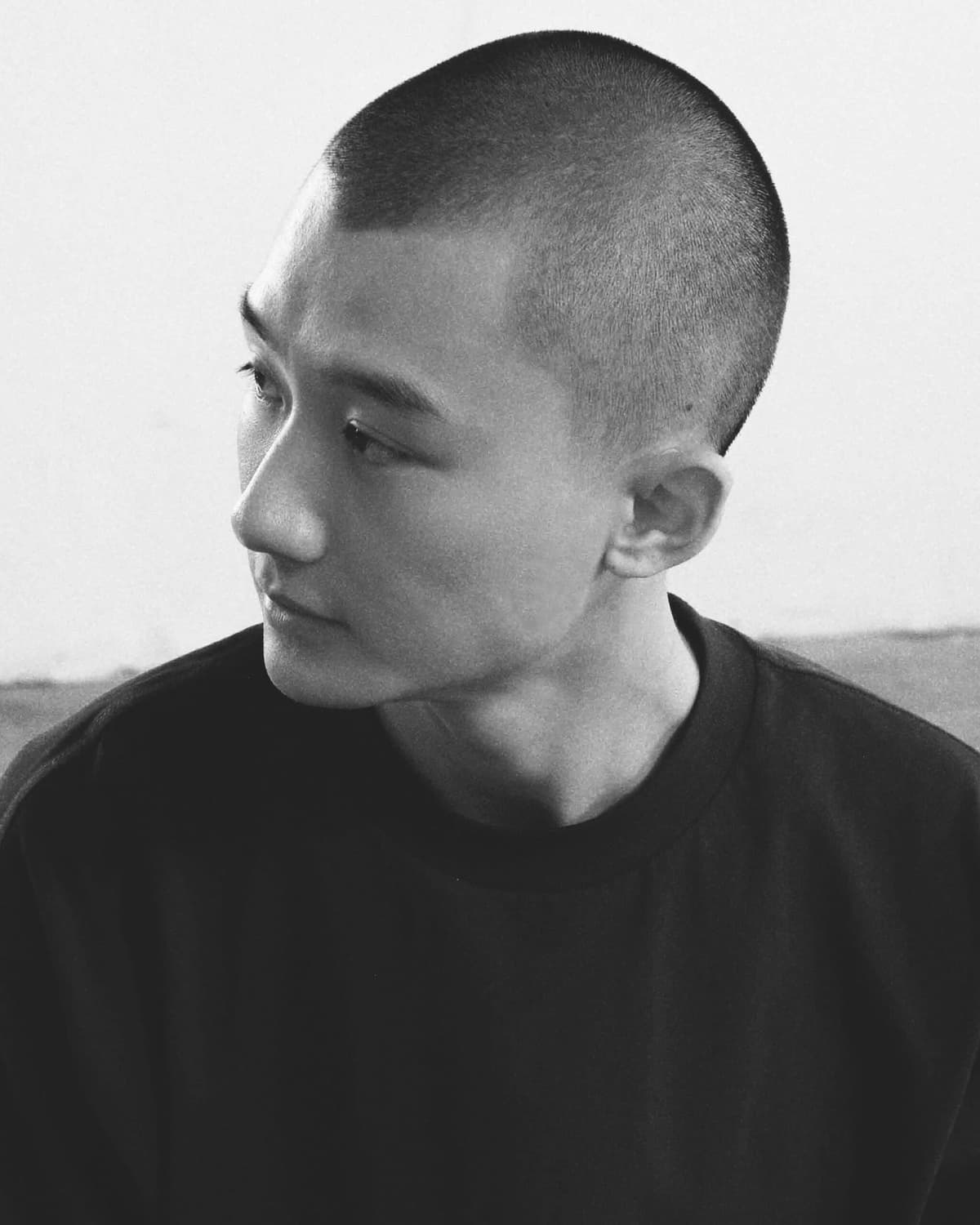 Black-and-white portrait of a man with a shaved head in a dark sweater, looking to the side against a pale background