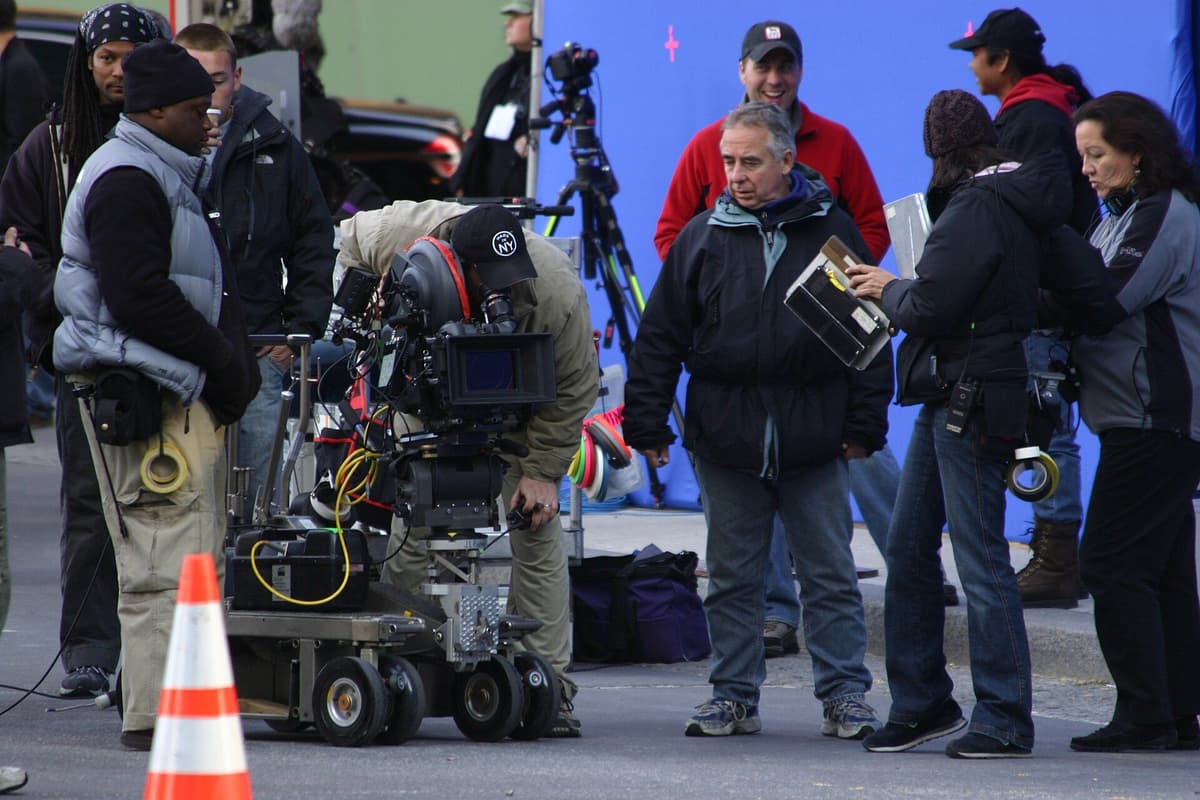 Film crew gathered around a camera on a wheeled dolly on a street, with a blue backdrop and orange traffic cone