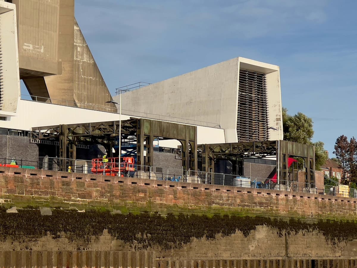 Concrete ventilation towers and steel structures beside a brick river wall, with workers and a lift platform on site.