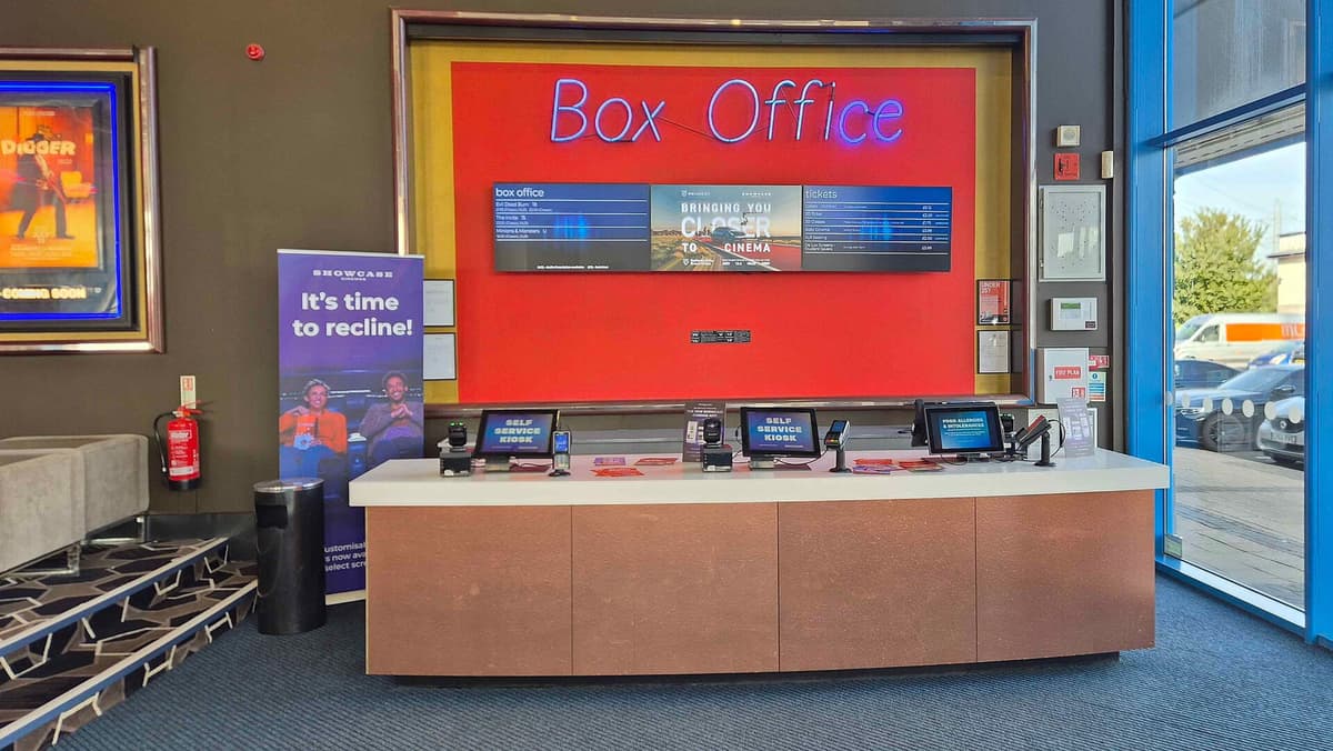 Cinema box office counter under a glowing neon sign, with self-service ticket kiosks and a red display wall in the lobby.