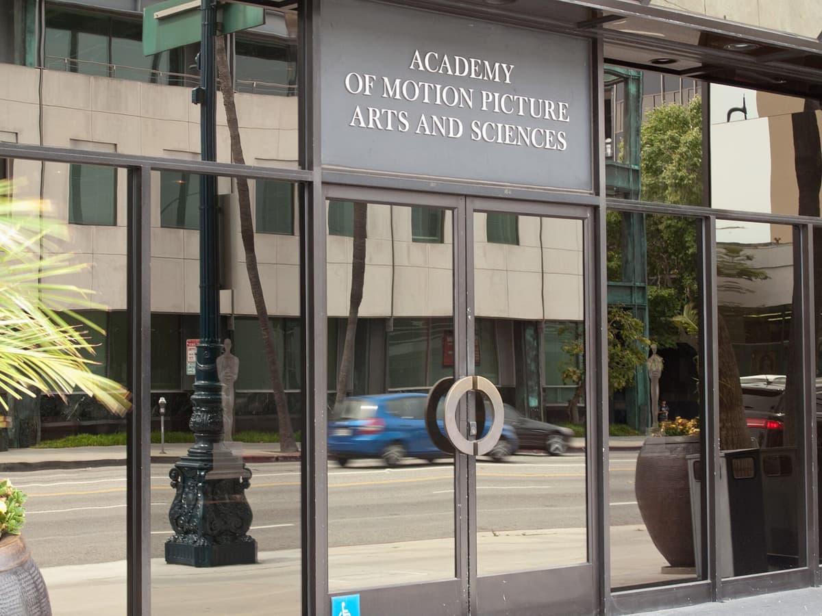 Glass entrance of the Academy of Motion Picture Arts and Sciences building, with a blue car passing on the street.