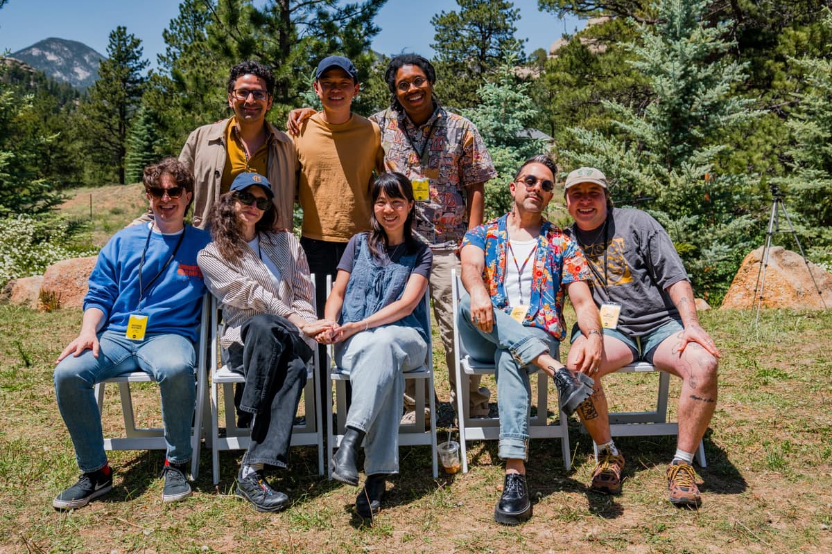 Eight people posing outdoors among pine trees and boulders, some seated on white chairs and some standing, smiling at the camera.