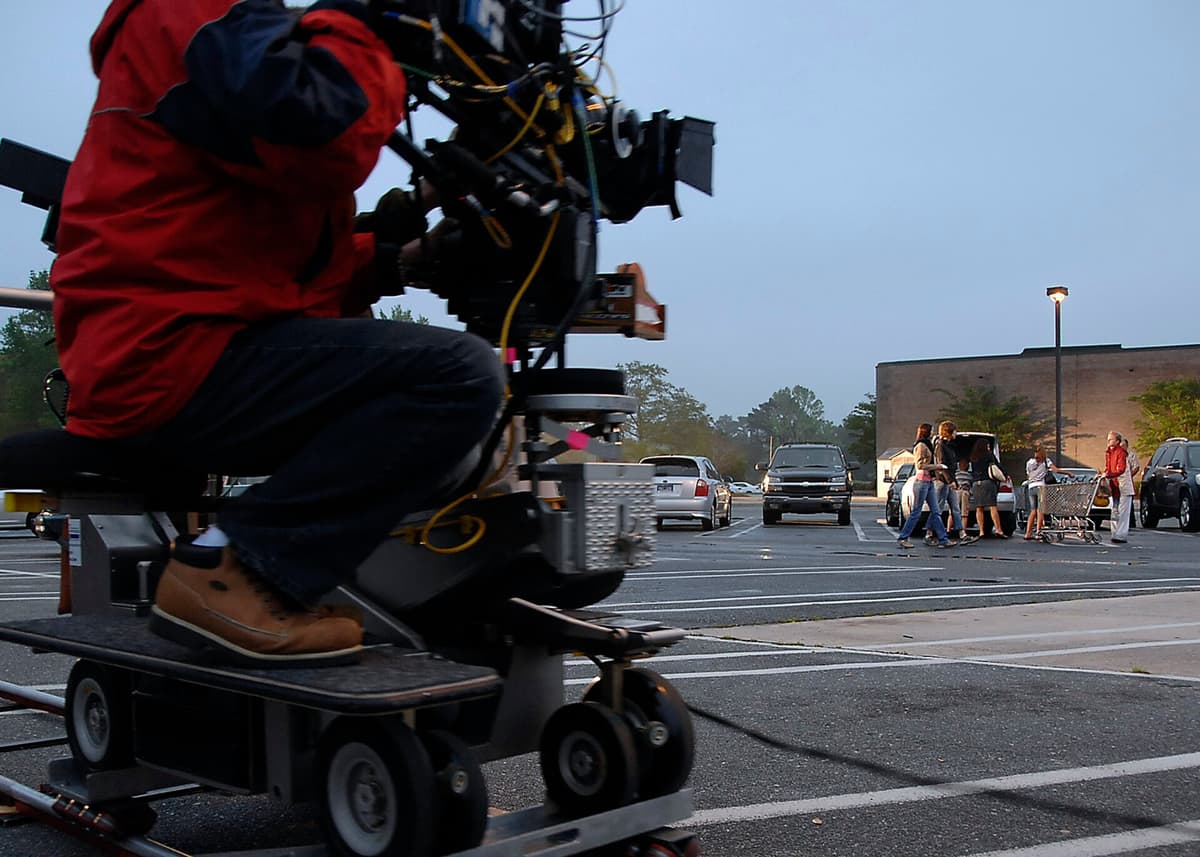 Camera operator in a red jacket on a dolly-mounted film camera rig, filming a scene in a parking lot at dusk