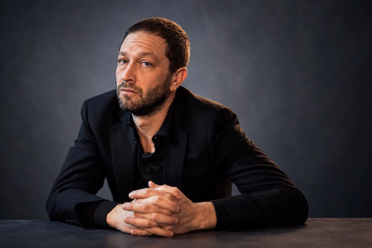 Ebon Moss-Bachrach in a black jacket, seated at a table with his hands clasped