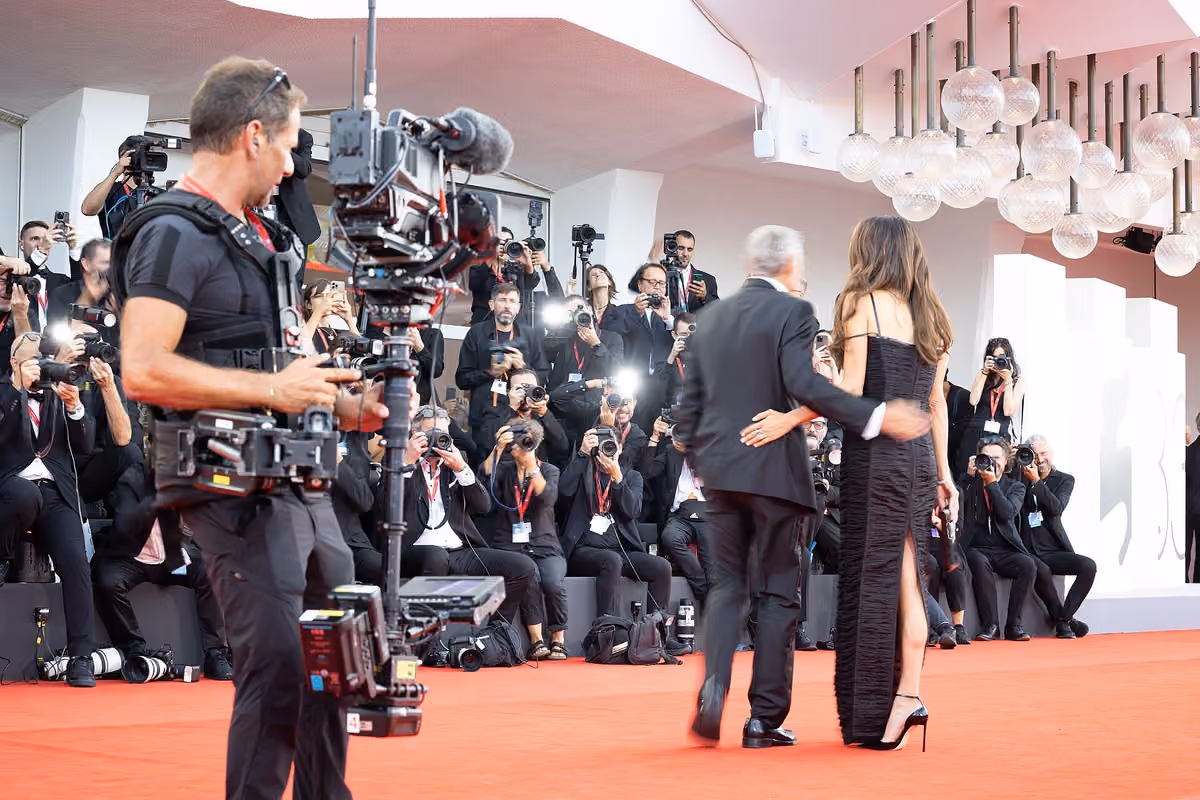 A couple walk down a red carpet past a dense crowd of photographers at a film festival