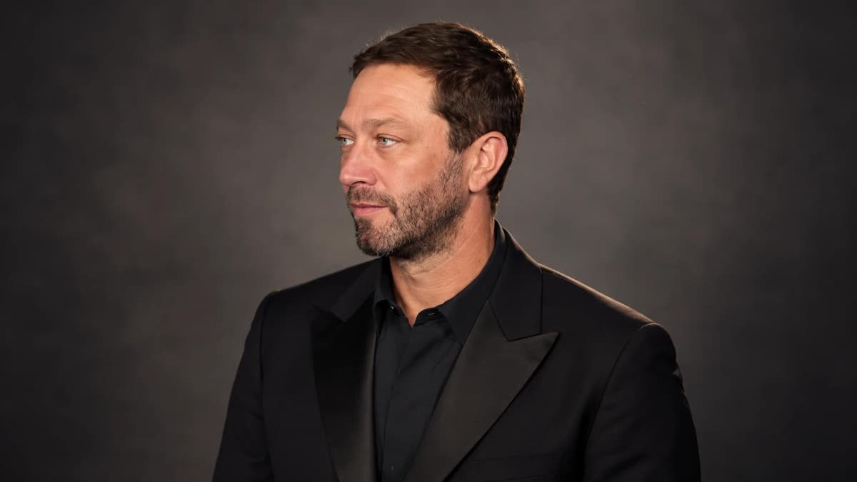 Ebon Moss-Bachrach in a black suit and shirt, looking off camera