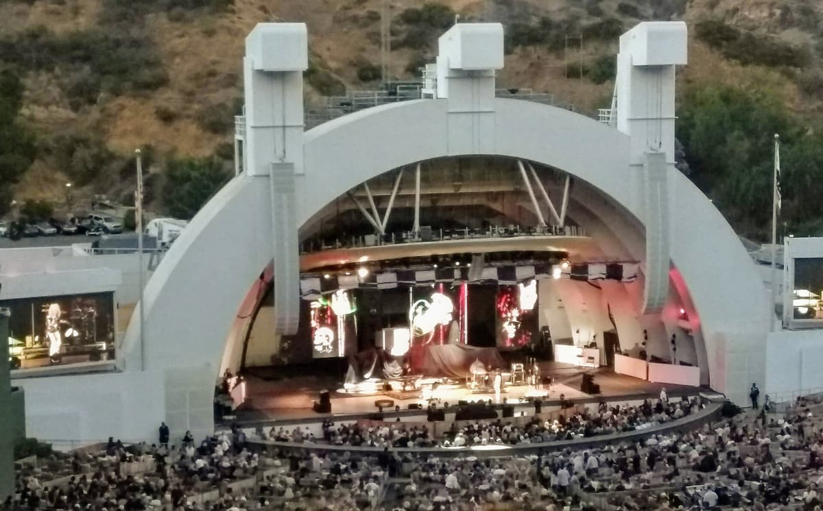 Outdoor concert in progress on a white arched stage, with a large crowd seated in the venue below.