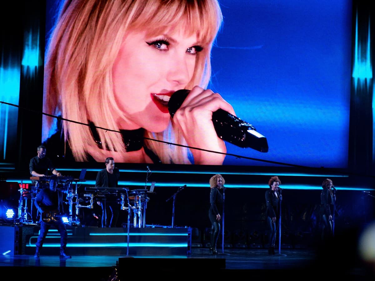 Singer on a large video screen holding a microphone, with a keyboardist, guitarist and three backing singers on stage