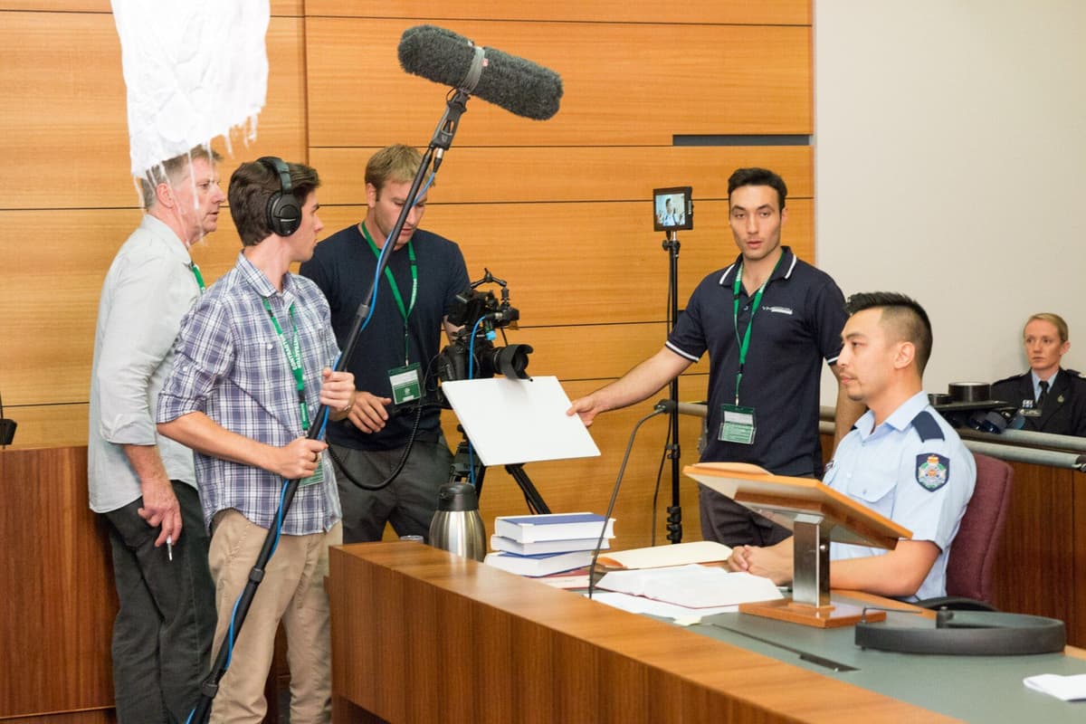 A film crew with a boom microphone, camera and monitor working on a set that looks like a courtroom