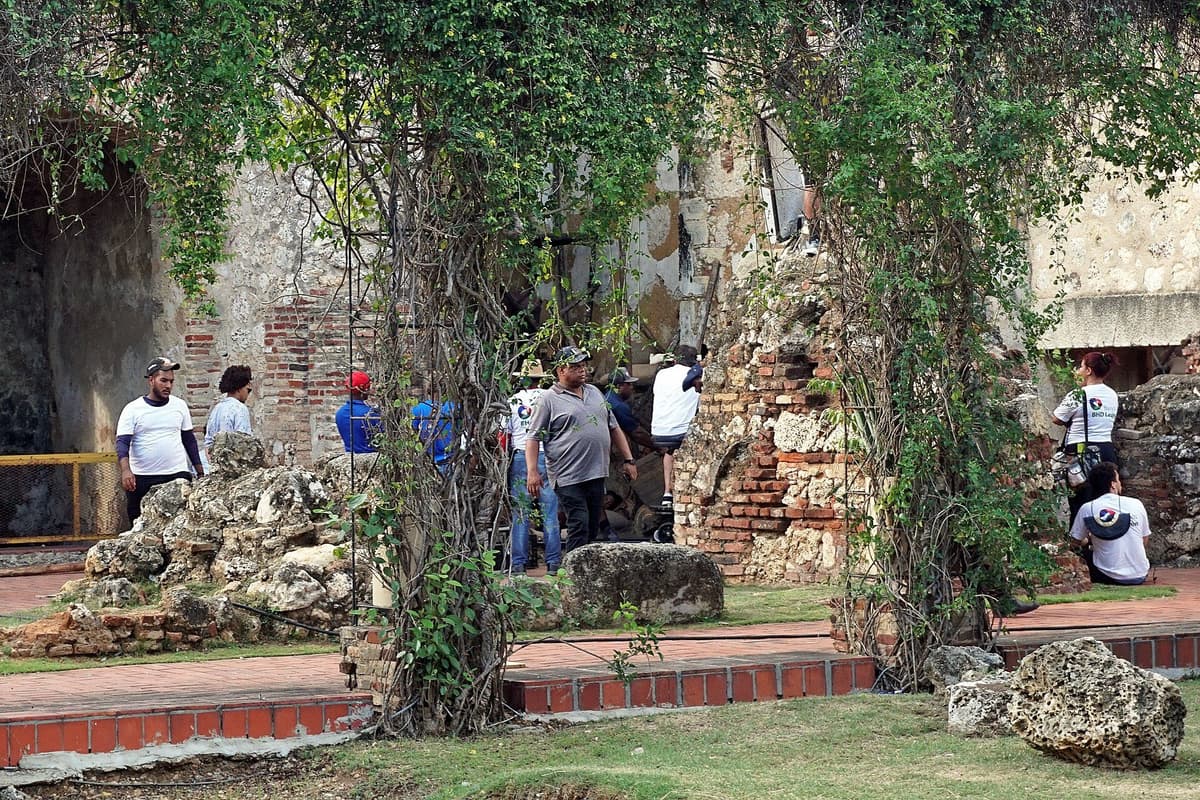 Crew members standing and sitting among stone and brick ruins framed by leafy vines