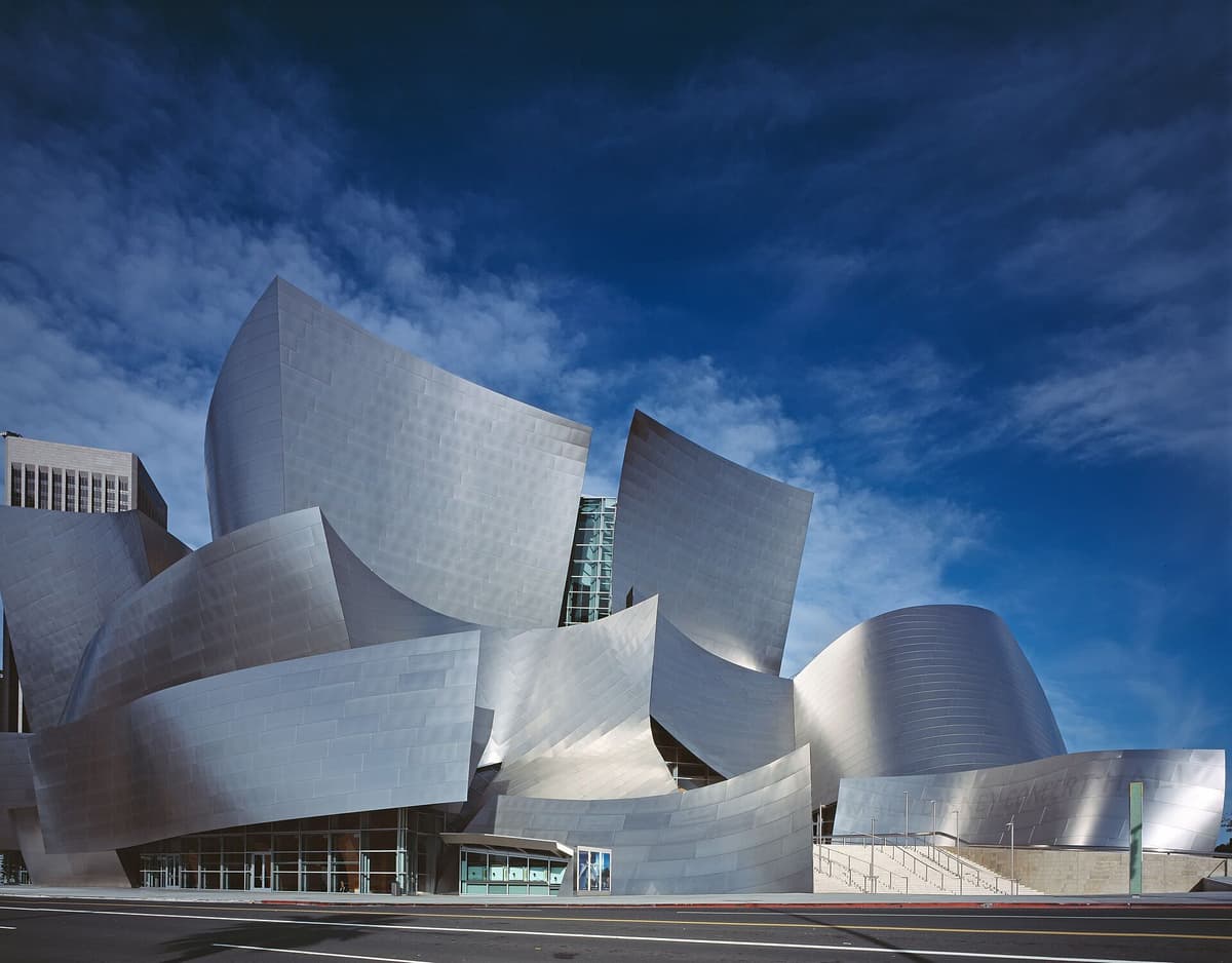 Exterior of Walt Disney Concert Hall with curving metallic panels against a blue sky, viewed from street level.